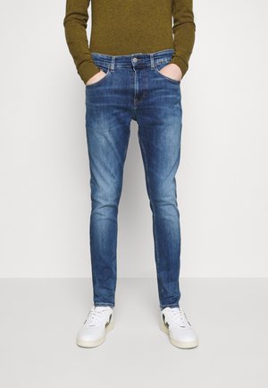 Jeans de mezclilla azul oscuro con un corte ajustado, bolsillos frontales y un acabado desgastado, combinados con zapatillas blancas y una camiseta texturizada de color verde oliva.
