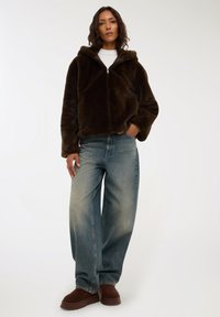 Felpa con zip in ecopelliccia marrone e cappuccio, indossata sopra jeans larghi blu chiaro e scarpe slip-on marroni scuri. Texture liscia e vestibilità rilassata.