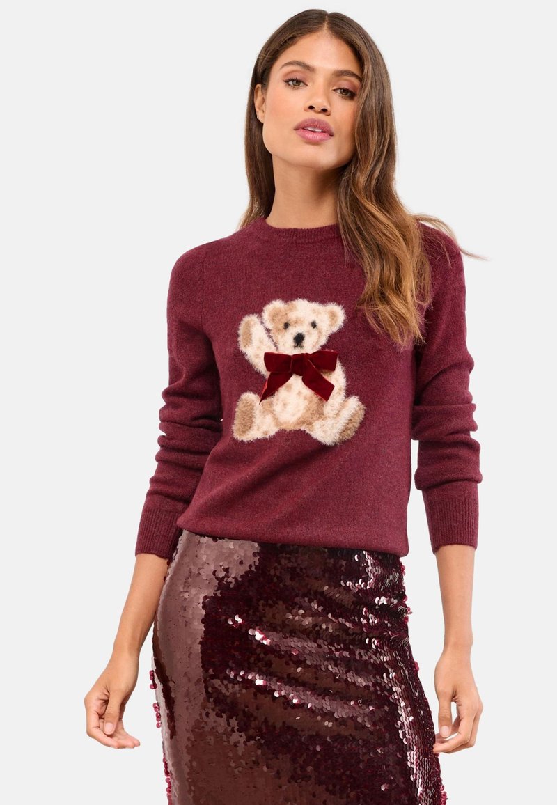 Vrouw draagt een bordeauxrode trui met een teddybeer- en strikontwerp, gecombineerd met een glanzende donkerrode pailletten rok.