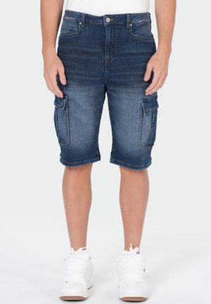 Homme portant un short cargo en jean bleu foncé et des baskets blanches, debout, les bras détendus le long du corps.