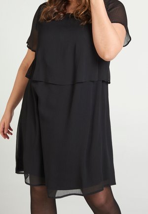Femme portant une robe noire superposée, longueur genou, à manches courtes, avec des détails en tissu transparent sur les manches et l'ourlet.