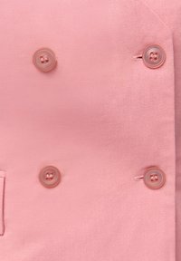 Světle růžový oblekový blejzr s texturovanou látkou, který má dva odpovídající knoflíky a jednu boční kapsu.
