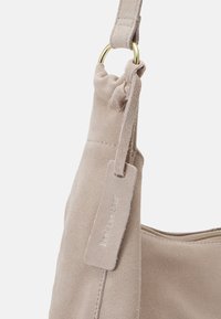 Beige Wildledertasche mit runder Form, ausgestattet mit einem goldenen Metallring und einem Stoffetikett, das mit "Echtleder" geprägt ist. Glatte Textur, lässiges Design.