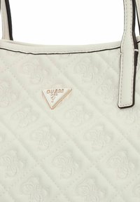 Borsa in finta pelle bianca trapuntata con logo dorato a triangolo "GUESS" e motivo logo sottile. Caratterizzata da bordi neri e manici doppi.