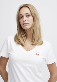 Weißes Baumwoll-T-Shirt mit V-Ausschnitt, das auf der linken Brustseite ein kleines, gesticktes Kirschdesign in Orange und Grün zeigt.