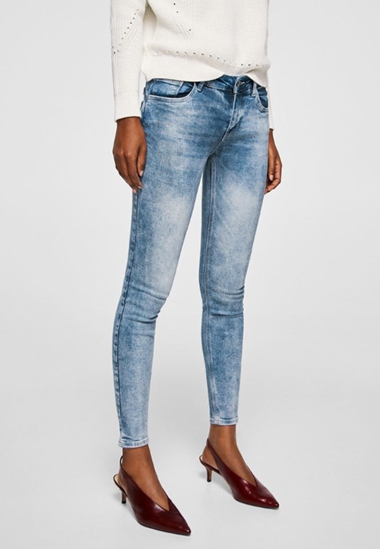 Femme portant un jean skinny bleu clair, un pull en tricot blanc et des escarpins slingback bordeaux à bout pointu, debout devant un fond uni.