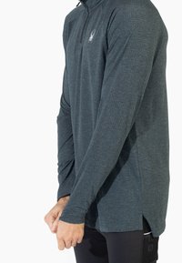 Maglia a maniche lunghe con zip nel collo, di colore grigio scuro, caratterizzata da un motivo testurizzato, fessure laterali e un logo a forma di ragno sul petto. Realizzata in tessuto traspirante.