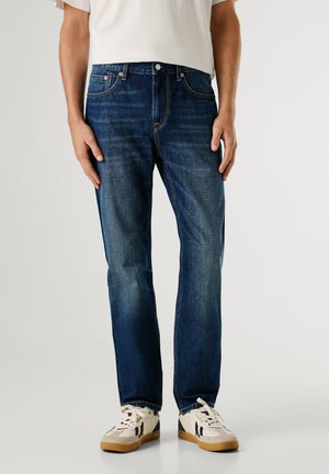 Dunkelblaue Jeans aus Denim mit geradem Schnitt, ausgestattet mit fünf Taschen, Kontrastnähten und einem Knopfverschluss, kombiniert mit weißen und beigefarbenen Sneakers.