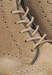 Beżowe zamszowe botki za kostkę z wiązaniem, posiadające perforowane akcenty oraz kontrastującą czarną krawędź wzdłuż oczek.