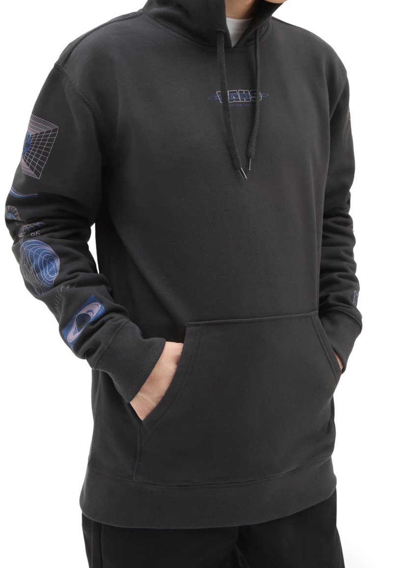 Personne portant un sweat à capuche noir avec des motifs sur le thème de l'espace sur les manches et la poitrine, les mains dans la poche avant, sur un fond blanc.