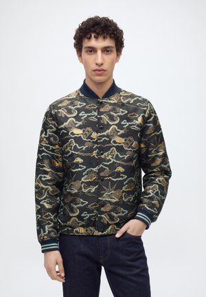 Jeune homme aux cheveux bouclés portant une veste bomber noire à motifs avec des dessins de nuages dorés et bleus et de dragons, debout sur fond blanc.
