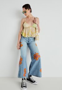 Calças largas de ganga azul clara com padrão floral e designs de hibiscos laranja, combinadas com um top cropped com padrões verde e amarelo e sapatilhas pretas.