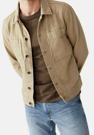 Man wearing light brown buttoned jacket and dark brown shirt paired with blue jeans, hands behind back, cropped at neck.