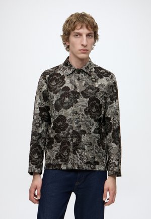 Ung person med krøllet hår iført en knapjakke med sort og gråt blomstermønster og mørkeblå jeans mod en hvid baggrund.