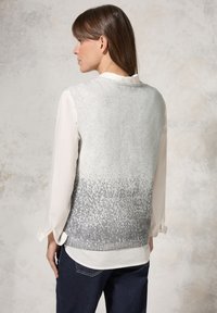 Chaleco de punto gris claro con una sección superior texturizada que se desvanece a gris oscuro en la parte inferior; superpuesto a una blusa blanca con mangas arremangadas.