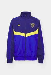 Modrá a žlutá sportovní bunda Adidas s vysokým límcem, zipovým uzávěrem a ribovanými manžetami. Obsahuje logo a tři pruhy na rukávech. Lehká tkanina.