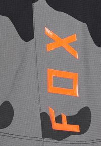 Närbild av grått tyg med svart camouflage-mönster och klara orangea bokstäver "FOX" sydda vertikalt.