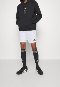 Persona vestita con un felpa nera con cappuccio, pantaloni corti bianchi Adidas, calze nere Adidas al ginocchio e scarpe da calcio nere Adidas, in piedi contro un muro neutro.