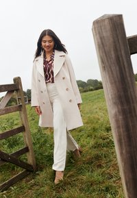 Podwójnie zapięty kremowy płaszcz z dużymi guzikami, noszony na wzorzystej bluzce i szerokich białych spodniach. Całość dopełniają brązowe botki na obcasie.