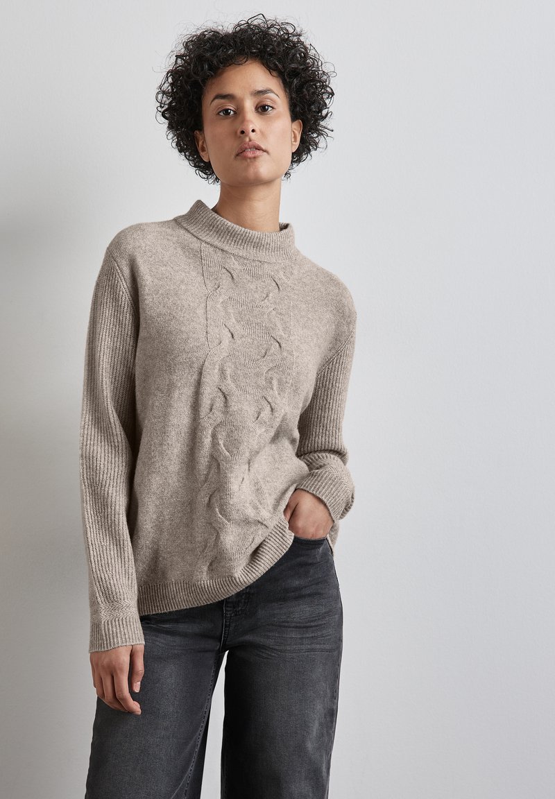 Maglione beige lavorato a maglia con colletto alto e motivo a trecce lungo il davanti, abbinato a pantaloni grigi scuri a gamba larga.
