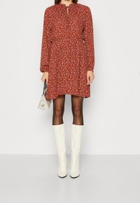Vestido largo de flores rojas con mangas largas y cintura fruncida, escote en forma de llave, combinado con botas de tacón alto blancas hasta la rodilla y un pequeño bolso blanco.