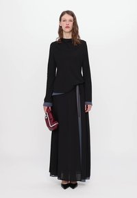 Black long-sleeve top with a draped silhouette, paired with a flowing black skirt featuring blue accents. Model holds a burgundy handbag.