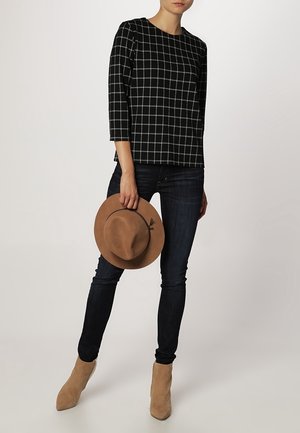 Žena v černo-bílém kostkovaném topu, tmavých úzkých džínách, hnědých botách, držící u boku hnědý klobouk s širokým okrajem.