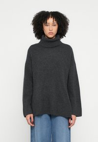 Pulover gri cu guler înalt, tricotat, cu croială lejeră, mâneci și tiv striate. Purtat cu blugi largi, de culoare albastru deschis. Textură netedă, design casual.