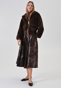 Braune Kunstfell-Bomberjacke mit Reißverschluss, kombiniert mit einem schwarzen Rock mit kupferfarbenem Blumenmuster und schwarzen Ballerinas.