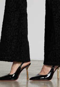 Zapatillas de salón negras de charol con punta afilada, con un tacón de aguja en tono dorado y pantalones negros texturizados de fondo.