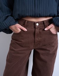 Άτομο που φορά καφέ παντελόνι ψηλόμεσο και μπλε cropped μπλούζα με ριγέ, με τα χέρια στις μπροστινές τσέπες.