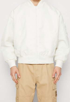 Personne portant une veste bomber blanche zippée avec des poignets élastiques et un pantalon cargo beige, debout devant un fond uni.
