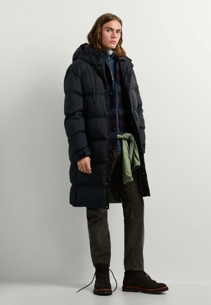 Jeune homme aux cheveux mi-longs portant un long manteau noir matelassé, une chemise à carreaux, un pantalon noir et des bottes noires à lacets, debout contre un mur uni.