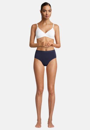 Weißer drahtloser BH mit verstellbaren Trägern und glatter Oberfläche, kombiniert mit hochgeschnittenen marineblauen Unterhosen. Das Model steht barfuß mit gefalteten Händen.