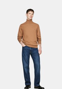 Brauner Rollkragenpullover aus Strickstoff, kombiniert mit blauen Jeans. Der Pullover hat ein kleines Logo auf der Brust und schmal geschnittene Bündchen.