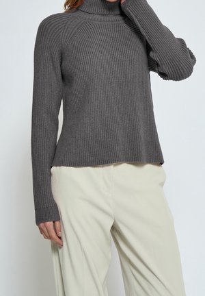Femme portant un pull à col roulé côtelé gris et un pantalon ample beige clair, posant avec une main près du visage.