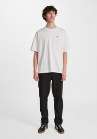 T-shirt bianco in cotone con colletto rotondo e maniche corte, con un piccolo logo nero sul petto; abbinato a pantaloni e scarpe nere.