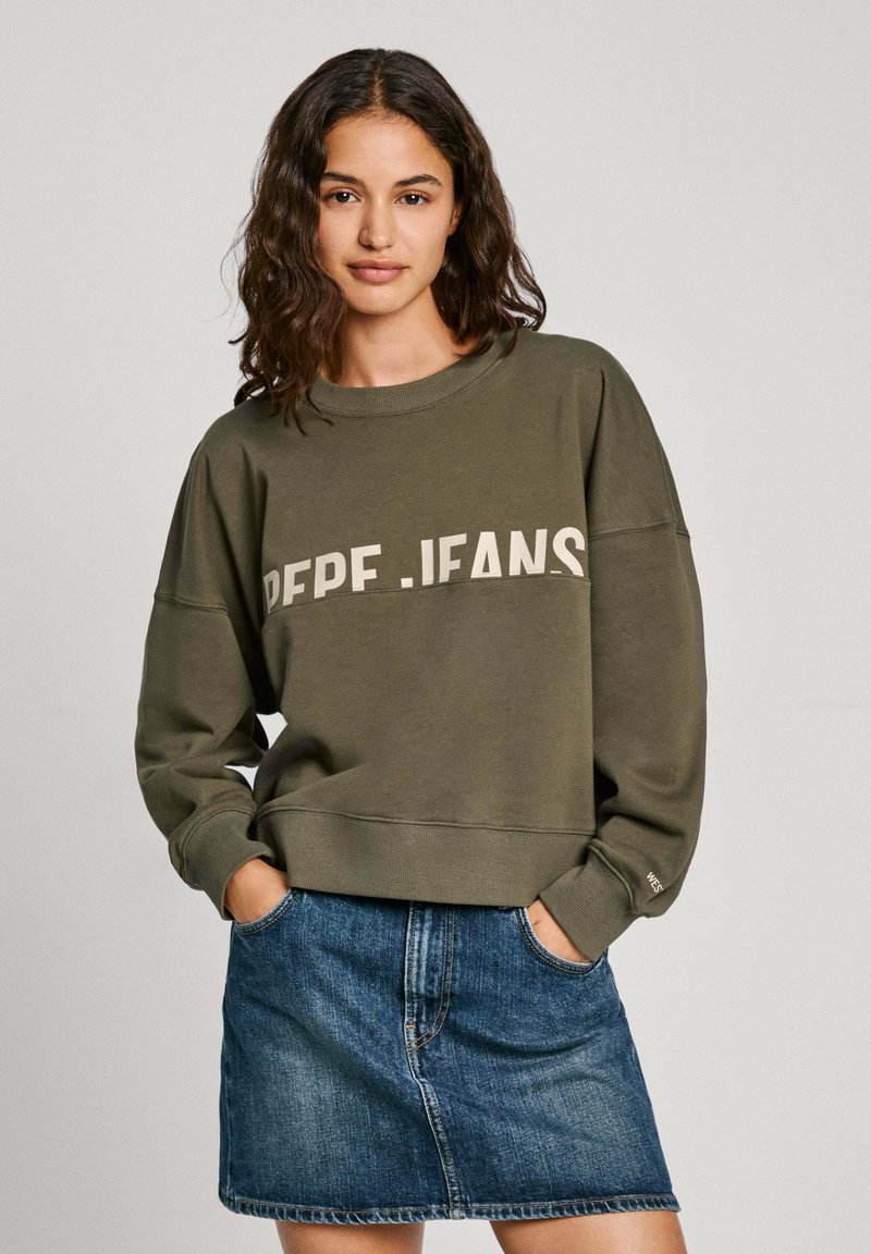 Sudadera verde oliva con el texto "PEPE.JEANS" en el pecho, cuello redondo y hombros caídos, combinada con una falda de mezclilla.