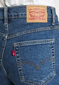 Pantalones vaqueros de mezclilla azul con una etiqueta de cuero marrón con detalles en rojo, diseño clásico de bolsillo trasero y costuras amarillas en contraste.