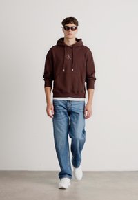 Brun bomull hettegenser med frontlomme og brodert logo, kombinert med lyseblå denimjeans og hvite joggesko. Modellen har på seg mørke solbriller.