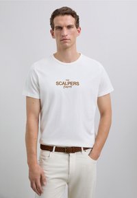 Uomo che indossa una maglietta bianca con la scritta "The Scalpers Company", pantaloni beige e una cintura intrecciata marrone, in piedi su uno sfondo semplice.
