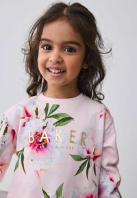 Heller rosa Sweatshirt mit bunten Blumenmustern und grünen Blättern, mit dem Schriftzug "BAKER" in goldener Schrift auf der Vorderseite. Weicher Stoff.
