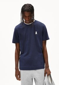 Donkerblauwe t-shirt met korte mouwen en een wit logo op de linkerborst, gemaakt van zachte stof, gecombineerd met grijze joggingbroeken.