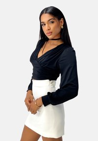 Blusa nera a maniche lunghe con scollo drappeggiato, abbinata a una mini gonna bianca aderente. Gli accessori includono un choker nero e orecchini dorati.