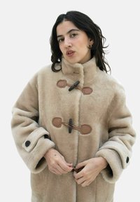 Beige fuskpälsjacka med stora manschetter och högt krage, med bruna snörstängningar och detaljer. Mjuk textur, oversize-design.