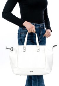 Person iført sort langærmet top og blå jeans, der holder en stor hvid læderhåndtaske i håndtagene i taljehøjde.