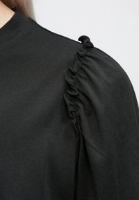 Fekete felső magas nyakkivágással és ráncolt fodor részletekkel a vállnál. Az anyag puhanak és simának tűnik. Egyszerű dizájn, látható minták nélkül.