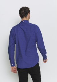 Camisa de manga larga azul con un pequeño patrón geométrico blanco, con cuello y puños abotonados, fabricada en un tejido suave.