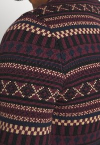 Pull tricoté avec une base bordeaux, présentant des motifs géométriques en noir, beige et bleu marine. Texture douce et coupe ajustée.