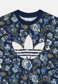 Tirkizna majica s cvjetnim uzorkom u mornarskoj boji s bijelim Adidas trefoil logotipom; sadrži šarene plave i žute cvjetove na teksturiranom materijalu.